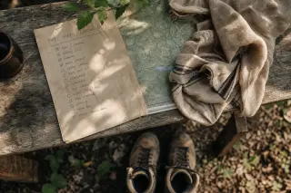 Table with handwritten checklist, old map, scarf, and worn boots in morning light, suggesting calm pre-adventure preparation outdoors.