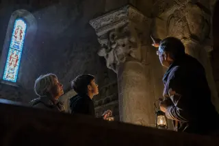 Intimate night tour inside a Corrèze church, with visitors quietly observing stone carvings by lantern light, soft moonlight filtering through stained-glass windows.