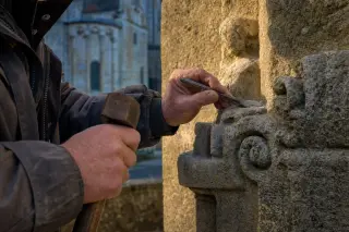 Elderly artisan carefully restoring stonework of the collégiale Saint-Martin d'Ussel, hands and tools highlighted by gentle afternoon light, with blurred historic façade in the background.