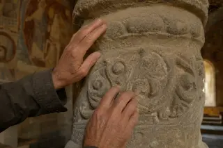 Elderly artisan's hands tracing Romanesque stone carvings in a softly lit historic chapel, showing texture and detail in gentle afternoon light.